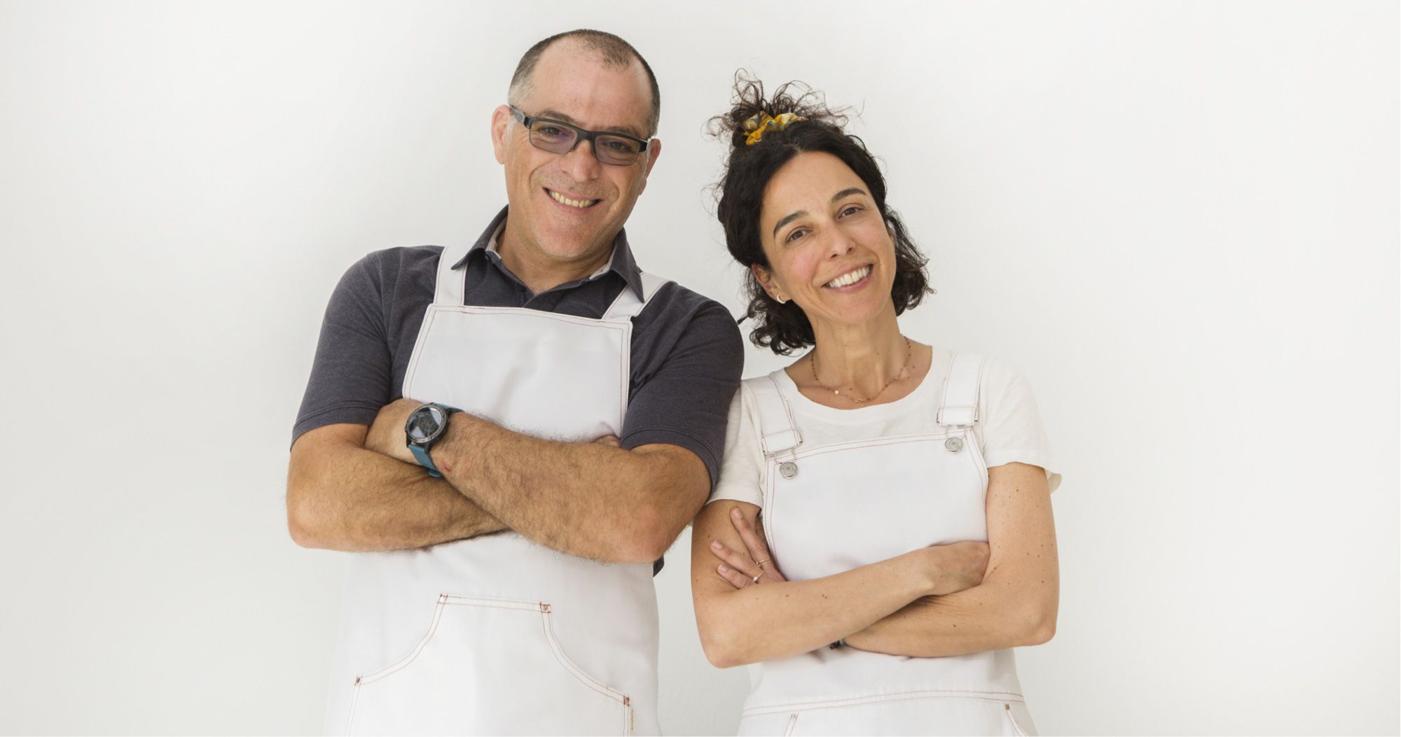 Two ice-cream makers in white aprons standing side by side against a plain pale wall, arms folded