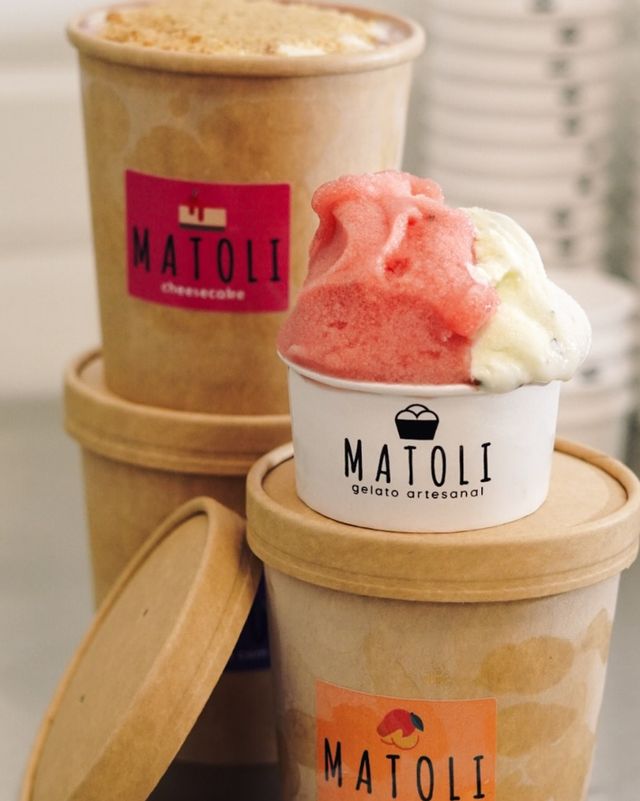 A white Matoli cup of two scoops, pink sorbet beside pale green, resting on kraft take-home tubs labelled with the Matoli mark