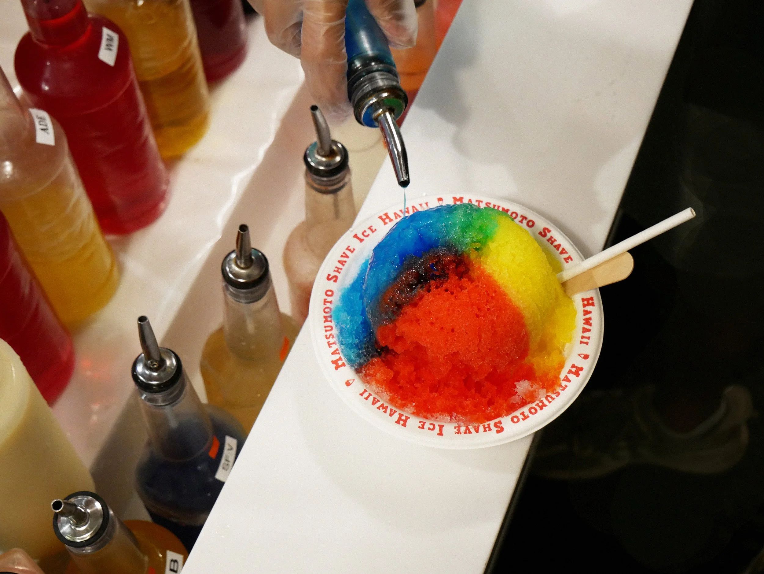 A bowl of rainbow shave ice beside a row of coloured syrup bottles