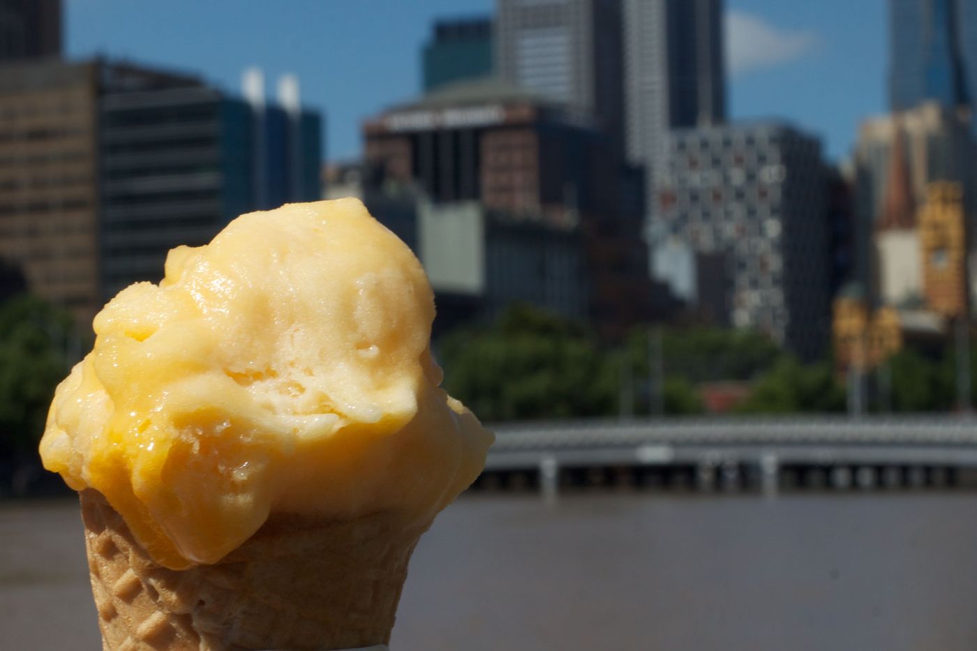 A scoop of yellow sorbet on a cone held up against the Melbourne skyline across the river