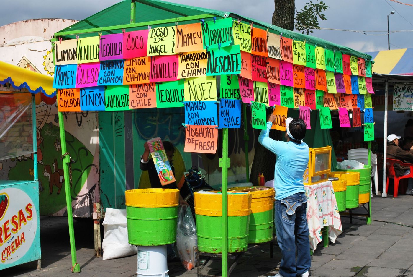 A nieve stall hung with hand-lettered flavour cards in pink, yellow and green, its vendor standing among stacked tubs