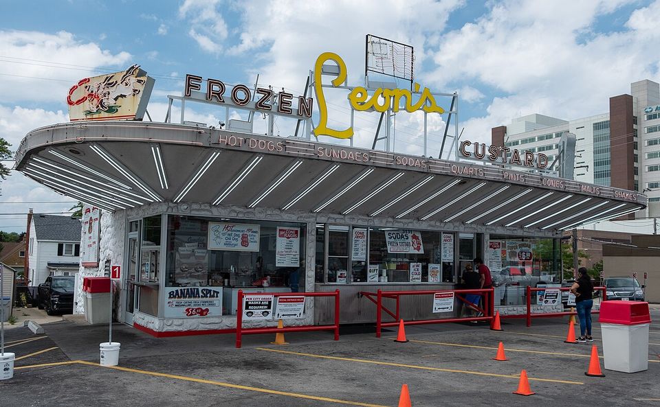 A drive-in under a long swooping canopy lettered FROZEN Leon's CUSTARD, with HOT DOGS, SUNDAES, CONES and MALTS spelled out along its edge
