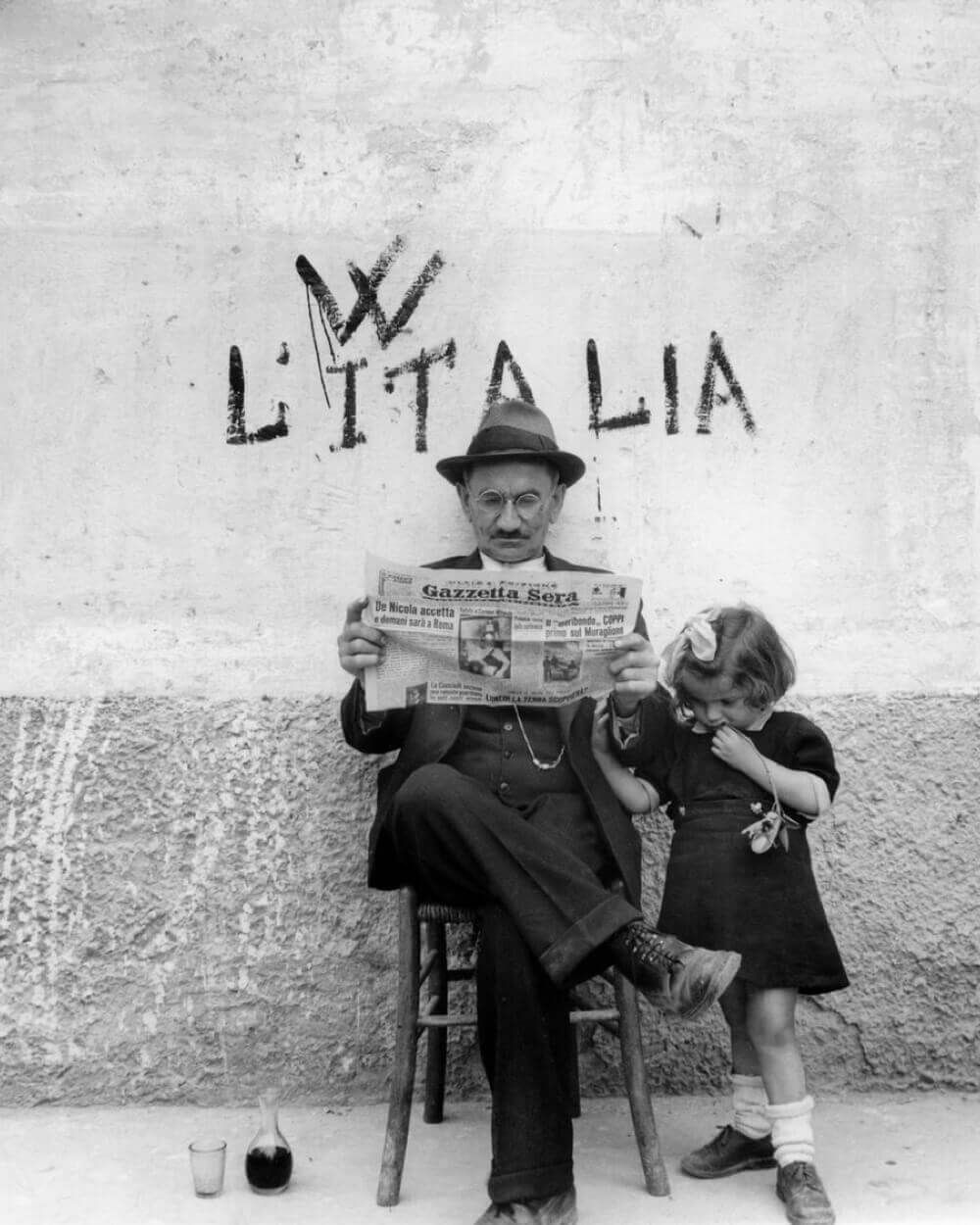 A black and white archive photograph of a man reading Gazzetta Sera beside a small child, against a wall painted L'ITALIA