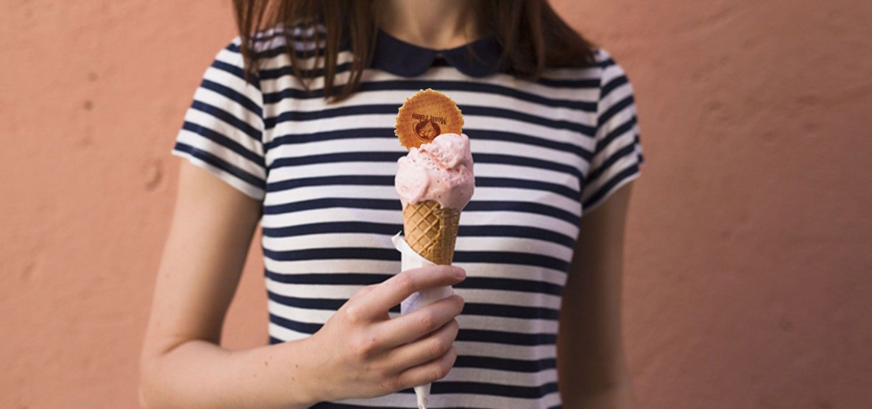 A cone of pale pink gelato held at chest height, the person behind it in a striped shirt