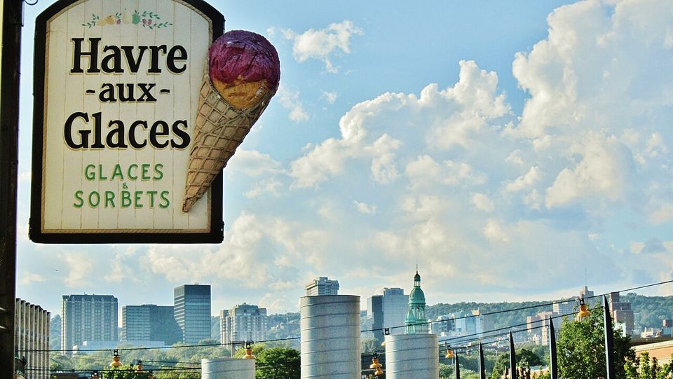 A hanging sign lettered Havre aux Glaces, GLACES & SORBETS, a moulded cone of purple sorbet fixed to it, the city skyline low behind