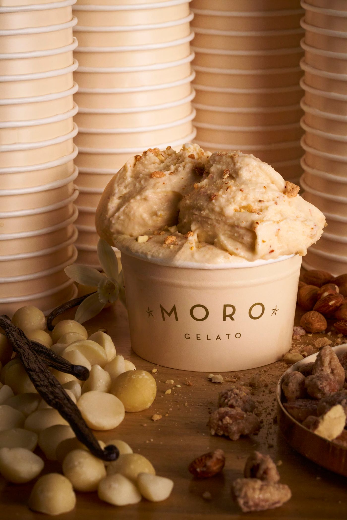 A cream paper tub printed Moro Gelato holds a scoop of pale nut gelato scattered with crumbled praline, set on wood among vanilla pods, white chocolate buttons, macadamia nuts and caramelised almonds, with stacks of empty tubs behind