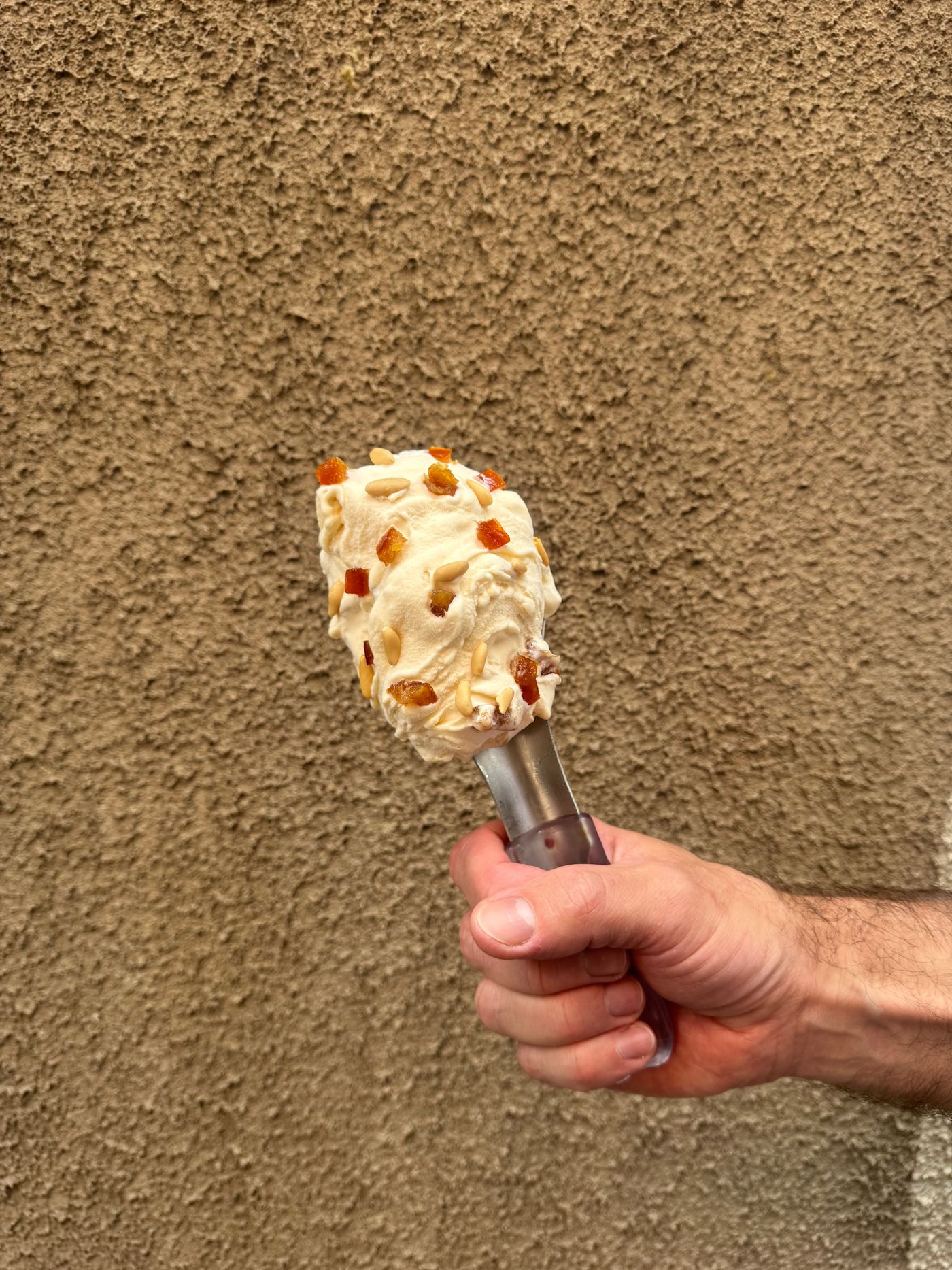 A hand holds a metal ice-cream spade carrying a scoop of pale gelato studded with orange candied fruit and pine nuts, against a rough beige rendered wall