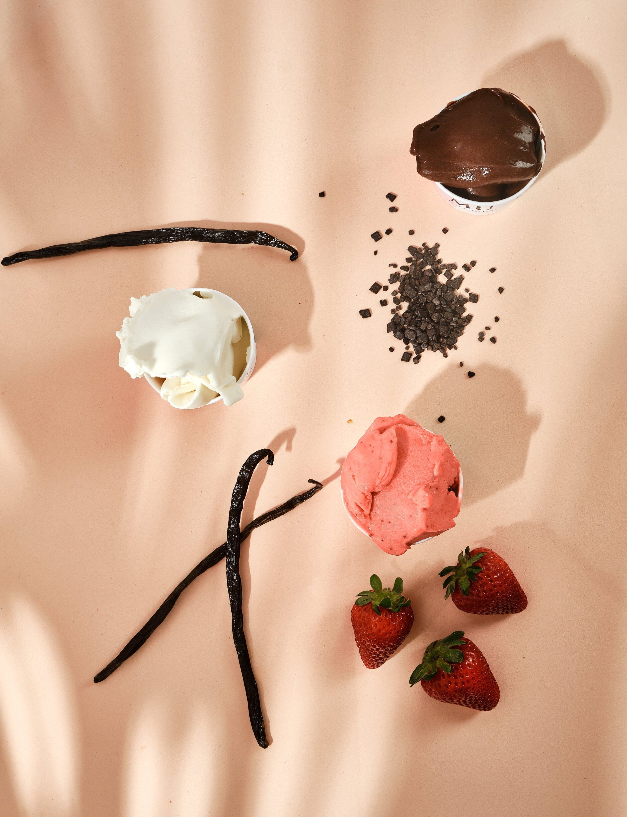 A flat lay of vanilla pods, fresh strawberries and small cups of vanilla, chocolate and strawberry gelato on a pink surface