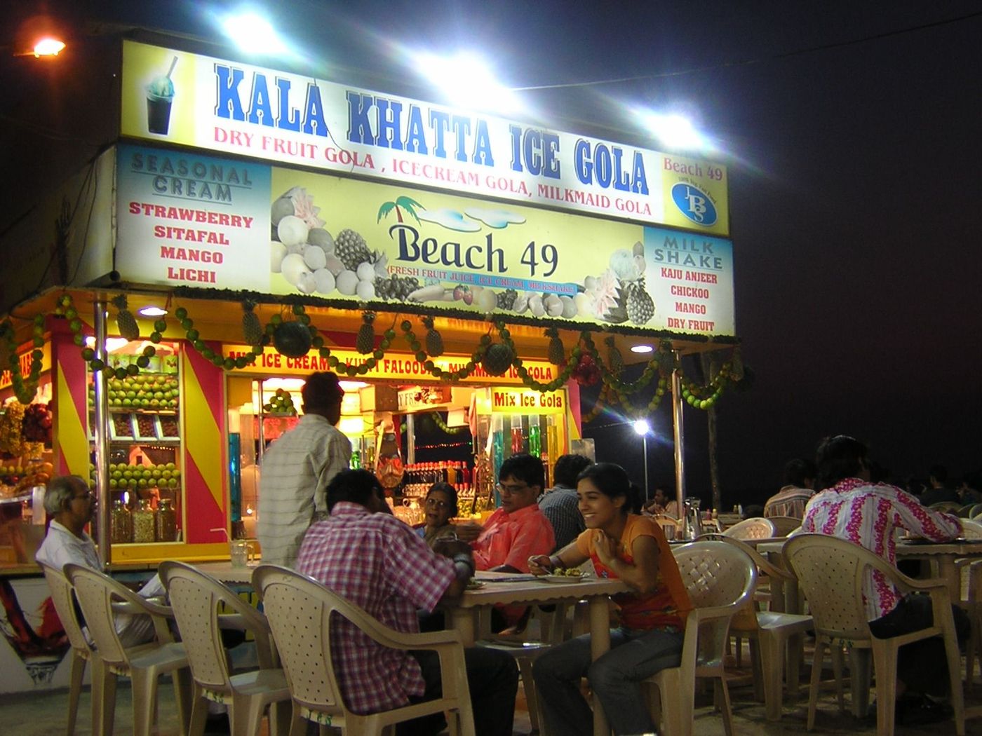 A lit stall on the beach at night signed KALA KHATTA ICE GOLA, people seated at plastic tables in front of it