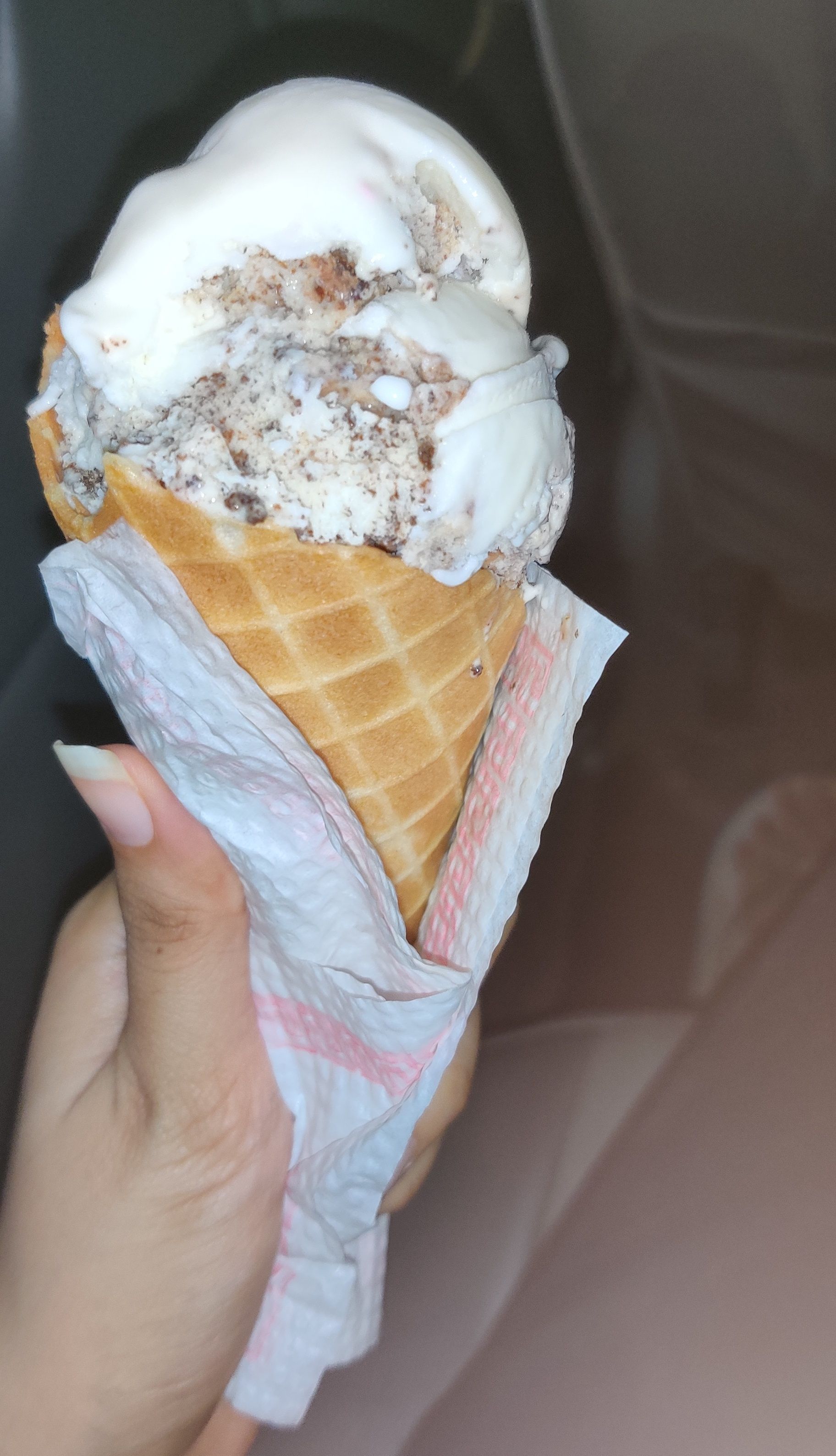 A double scoop of pale ice cream flecked with dark crumb, on a waffle cone held in a hand wrapped in a paper napkin