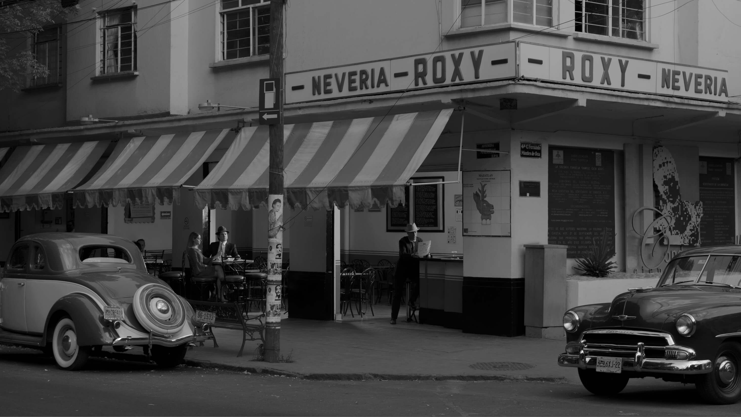 An archive black-and-white photograph of the Roxy shopfront with period cars at the kerb