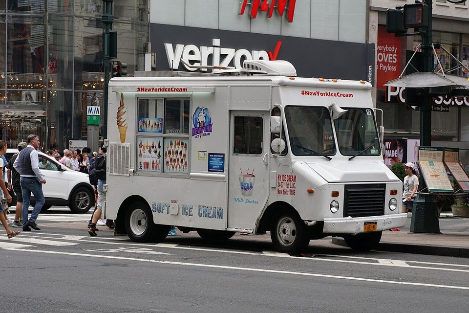 A white truck lettered SOFT ICE CREAM parked at a Manhattan crossing, rows of cones pictured on its window board