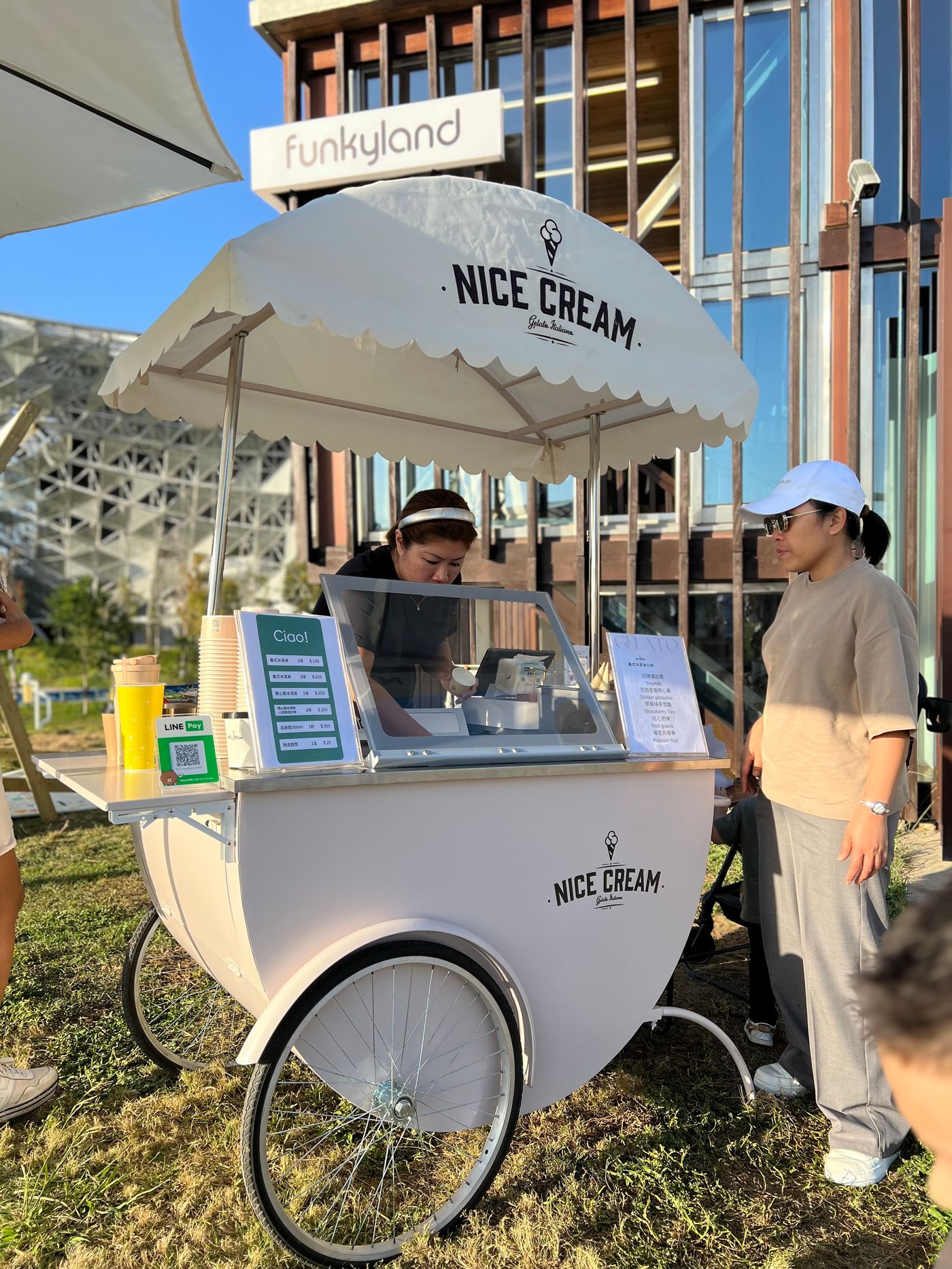 A white three-wheeled gelato cart with a scalloped canopy lettered Nice Cream — Gelato Italiano stands on grass, a server in black leaning over the open lid and a customer in a white cap waiting at the right