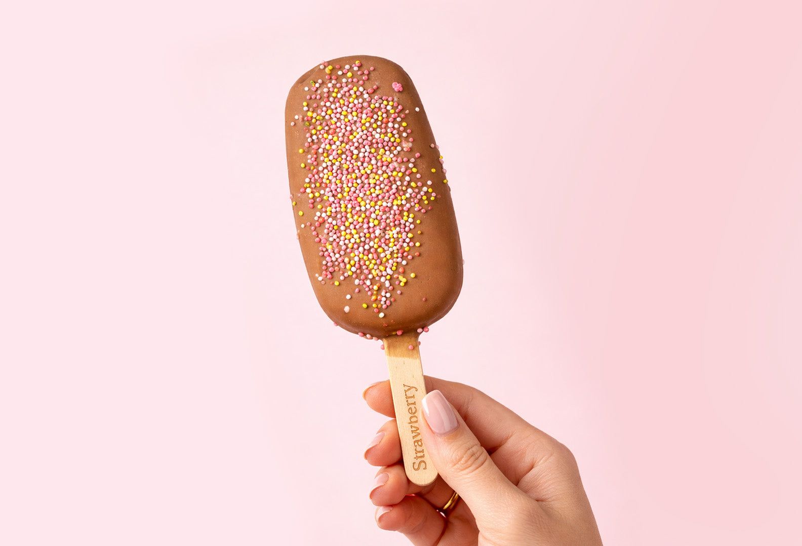 A hand holding a chocolate-coated ice lolly scattered with sprinkles, against pink