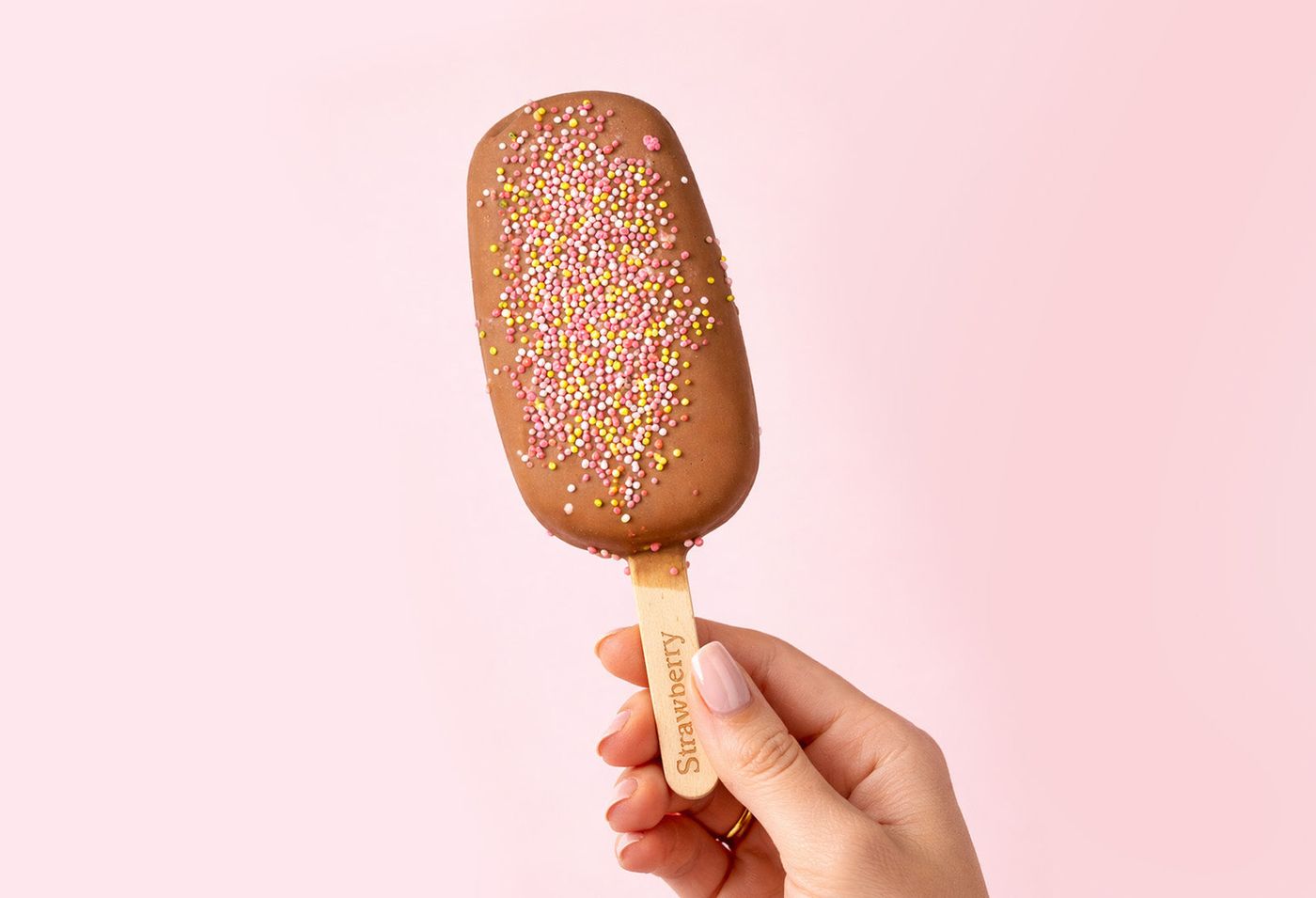 A hand holding a chocolate-coated ice lolly scattered with sprinkles, against pink