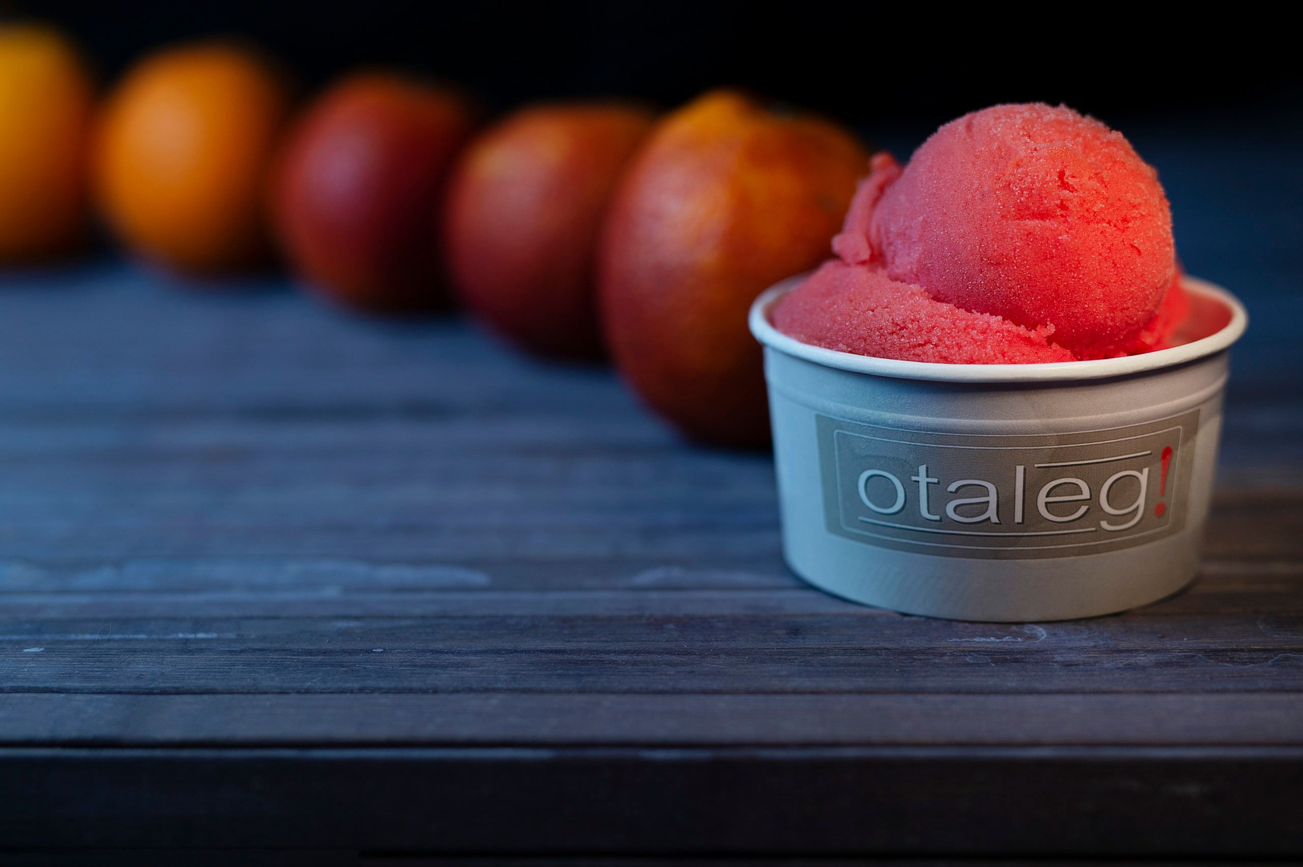 A scoop of red sorbet in a grey tub branded otaleg, on dark wood, with a row of blood oranges receding behind it