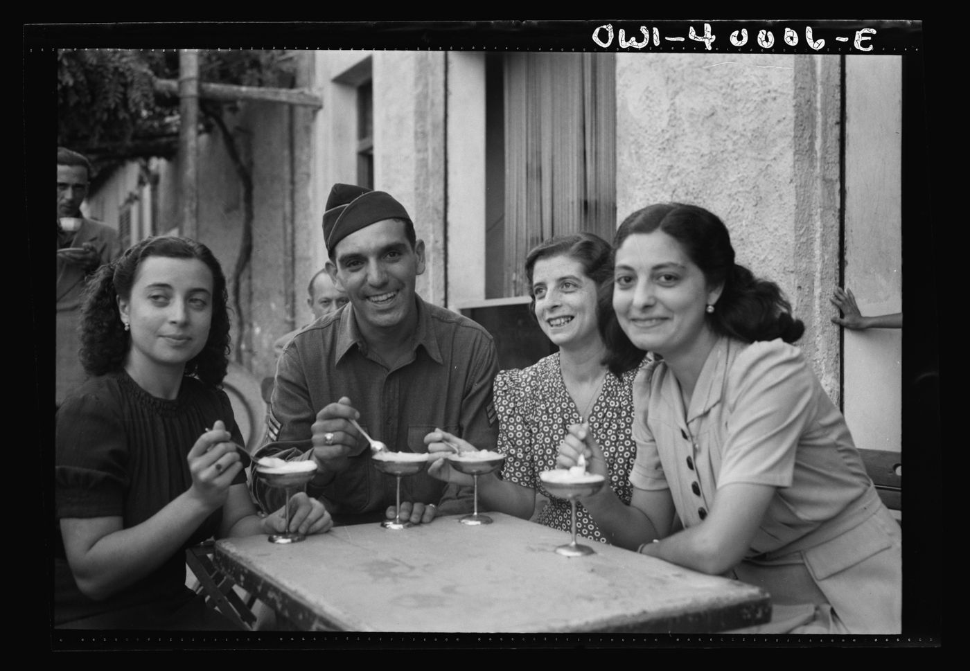 A 1943 photograph: an American soldier in uniform and three young women eating ice cream at a café table in Sicily