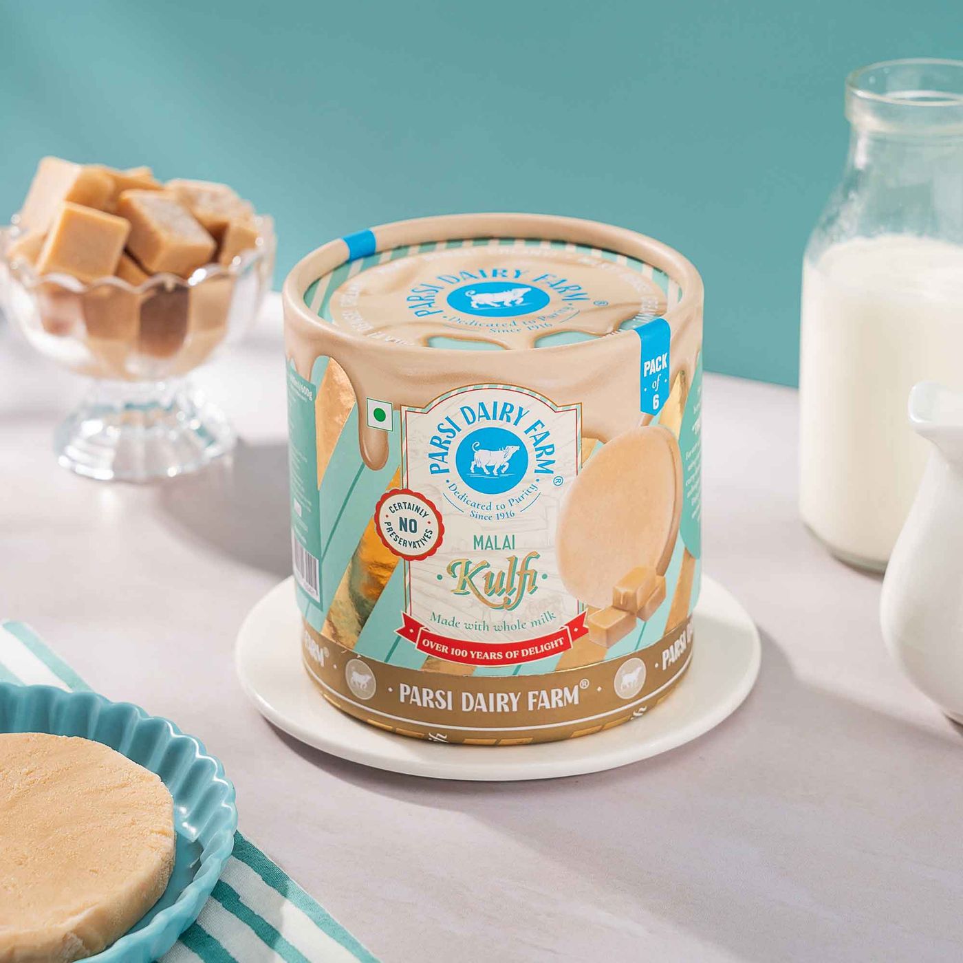 A Parsi Dairy Farm Malai Kulfi tub stands on a white saucer beside a glass bowl of kulfi cubes, a bottle and jug of milk, and a kulfi disc on a fluted blue plate