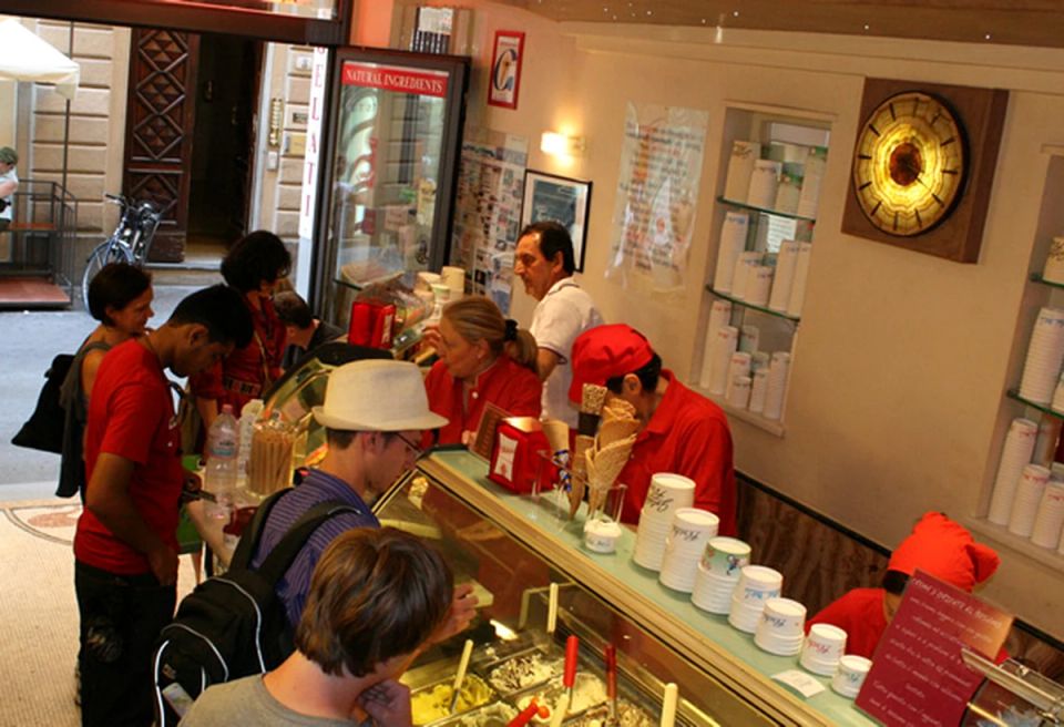 Staff in red shirts and red caps work behind a lit gelato case while customers lean over it, with stacked paper cups on shelves and a NATURAL INGREDIENTS sign by the door