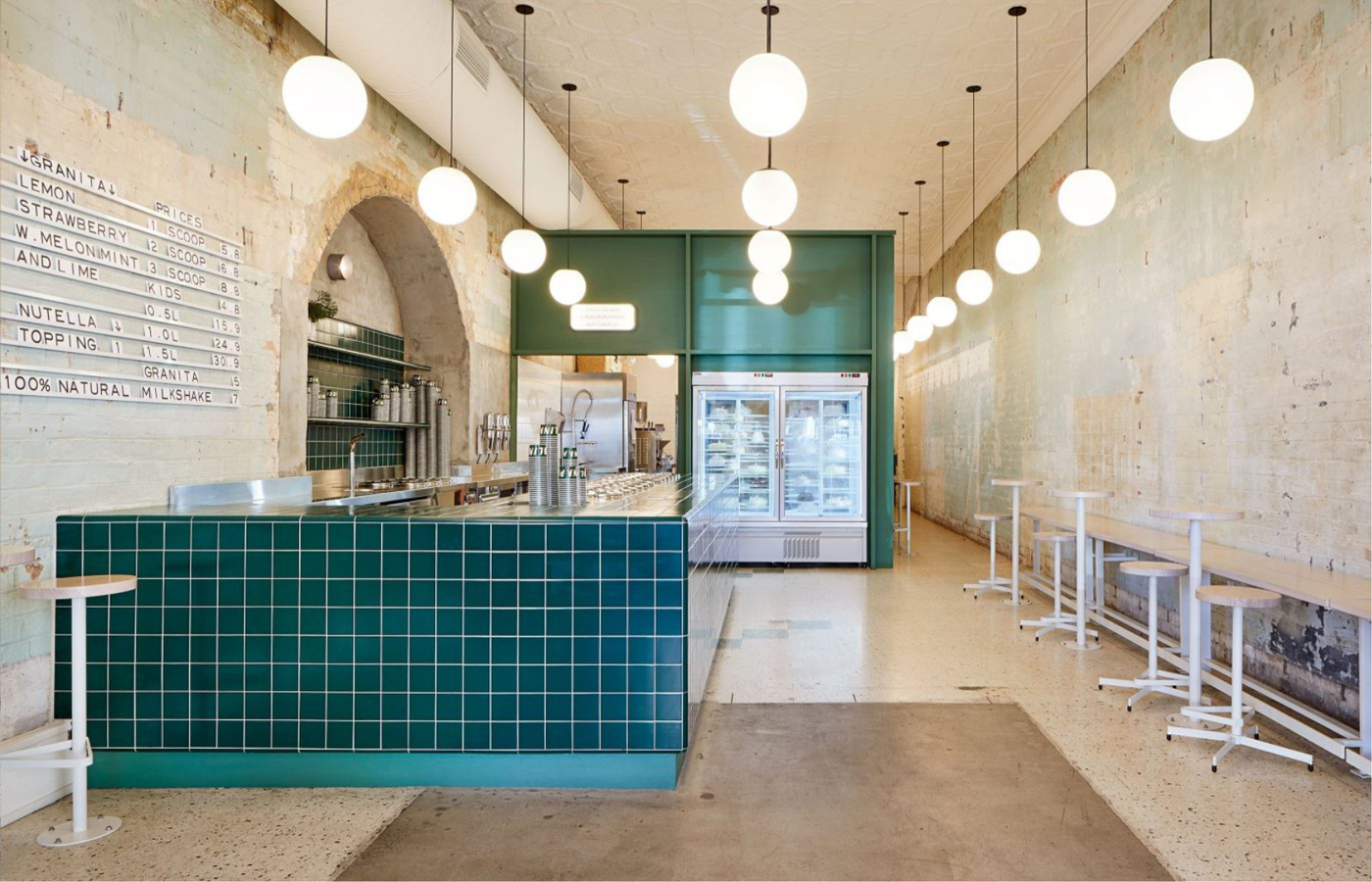 A teal-tiled counter runs down a narrow room under a row of white globe pendants, with a granita price board on the lime-washed wall, white stools along a bench, and a glass-door display fridge at the back