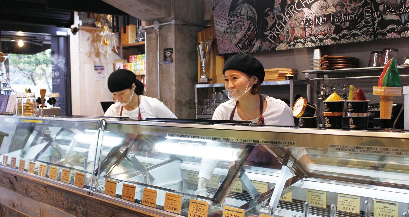 Two staff in black berets and white tops stand behind a long gelato case lined with bilingual flavour cards, under a chalkboard reading PREMARCHÉ — It's Not Fashion, But Revolution, with black tubs and display cones on the counter