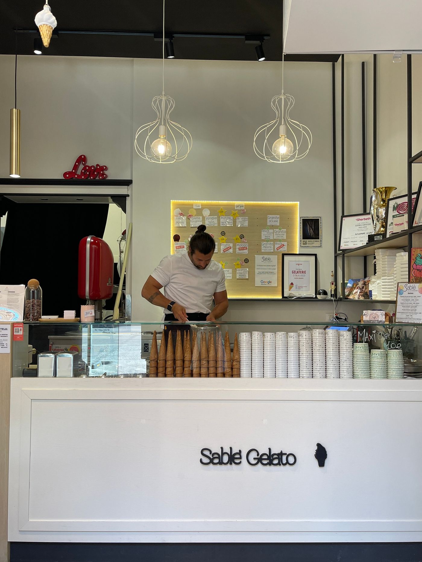 A man in a white t-shirt works behind a white counter lettered Sablè Gelato, with rows of waffle cones and stacks of paper cups on the counter and a lit handwritten flavour board and framed certificates on the wall behind