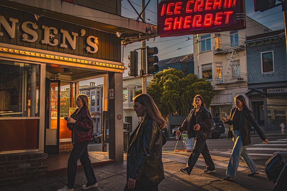 A neon ICE CREAM SHERBET sign burning red over a corner parlour at sunset, women crossing the street in front of it