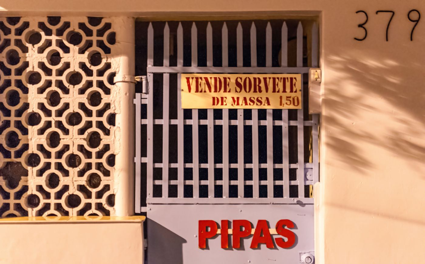 A hand-painted sign on a house gate reading VENDE SORVETE DE MASSA 1,50, and below it PIPAS