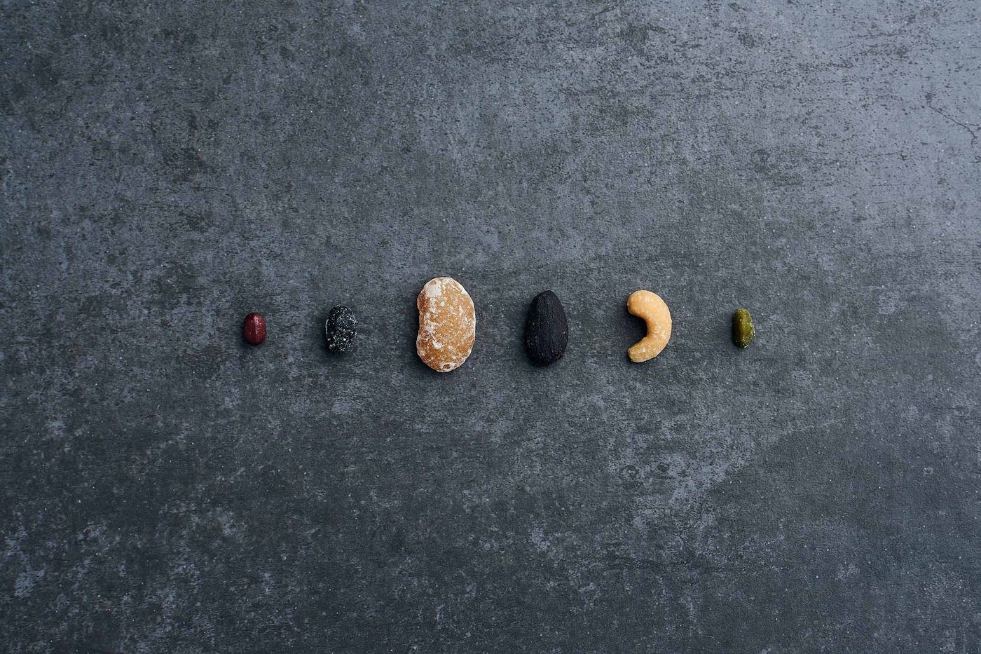 Six sugar- and chocolate-coated seeds and nuts — a small dark red bean, a black speckled nut, a large pale sugar-dusted oval, a black almond shape, a pale cashew and a small green kernel — lie in a row on mottled grey stone