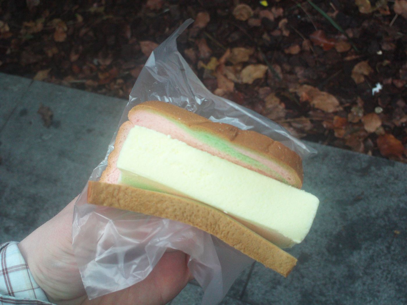 A slab of ice cream between two slices of bread, wrapped in plastic and held in one hand