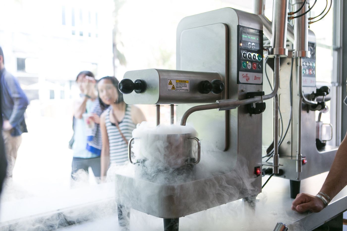 A stainless liquid-nitrogen machine branded Smitten Brrr spills white vapour over its bowl and across the floor while two customers watch from the far side of the shop window