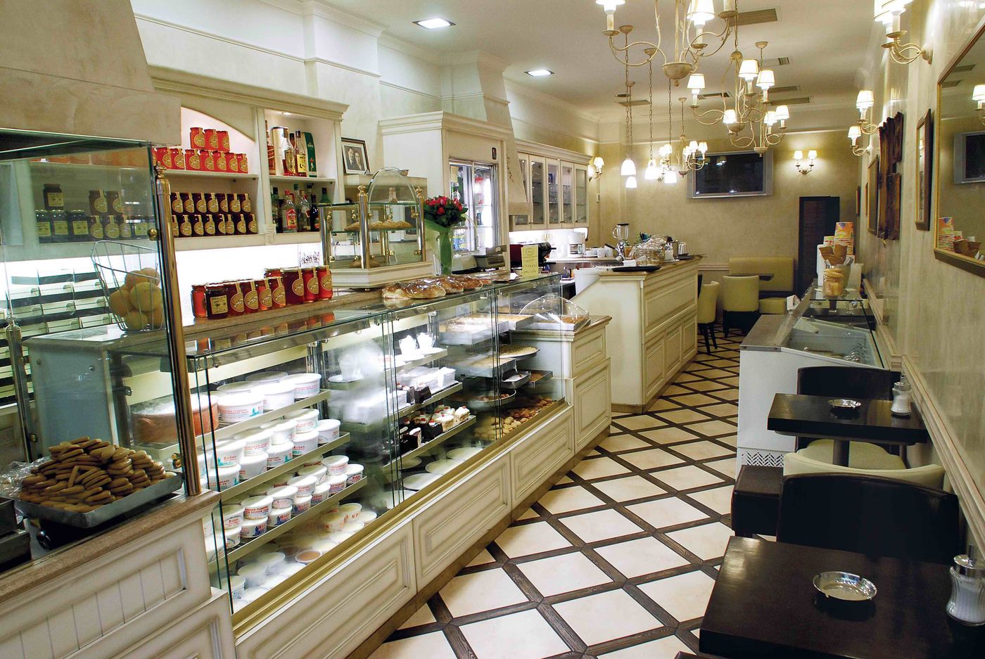 A long glass case holds tubs of yoghurt, trays of biscuits and small cakes, with jars of preserves and honey on the shelves above, chandeliers overhead and café tables on a diamond-tiled floor