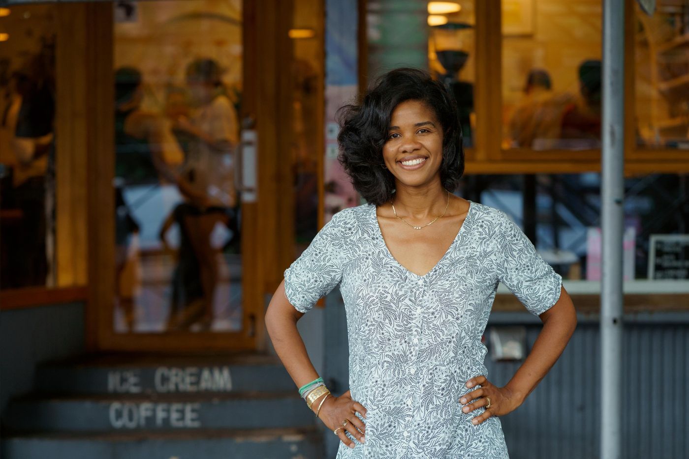 A woman stands with hands on hips outside the shop, in front of a wood-framed window and steps stencilled ICE CREAM / COFFEE