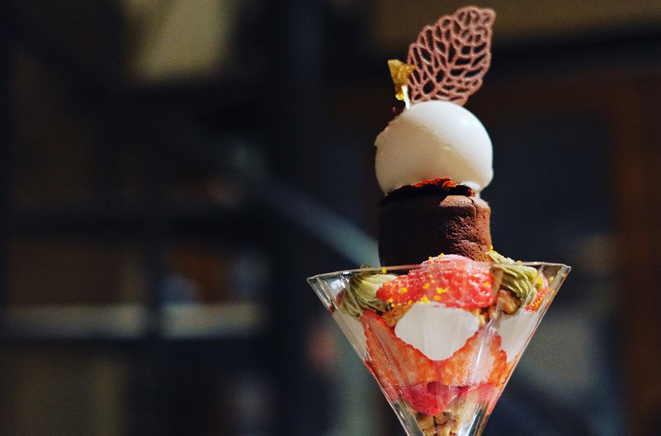A conical glass parfait holds strawberries, pistachio cream and crumble under a cocoa-dusted chocolate cylinder and a white gelato scoop topped with a chocolate lace tuile and gold leaf