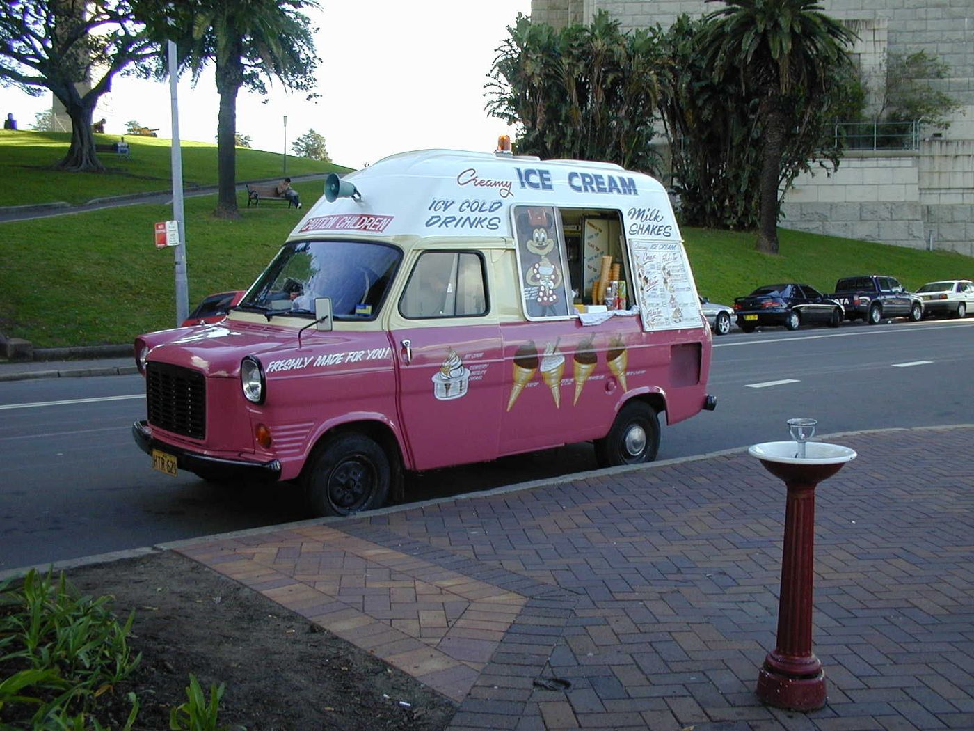 A pink and cream van lettered Creamy Ice Cream parked beside a park