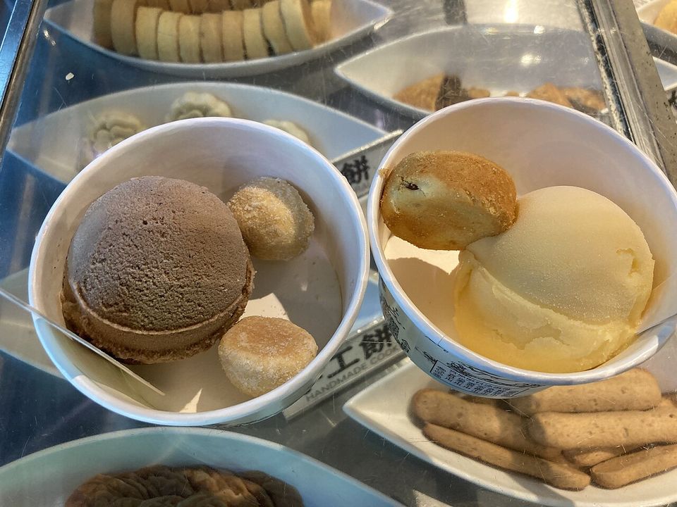 Two paper tubs of ice cream — one chocolate scoop pressed between wafers, one pale scoop beside a small pastry — set down on a glass case of biscuits