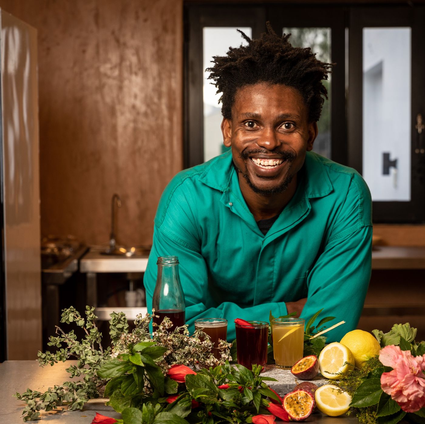 A man in a green shirt leans on a metal counter behind glasses of dark red, amber and yellow drinks, a bottle, sprigs of herbs, hibiscus flowers, halved lemons and passion fruit