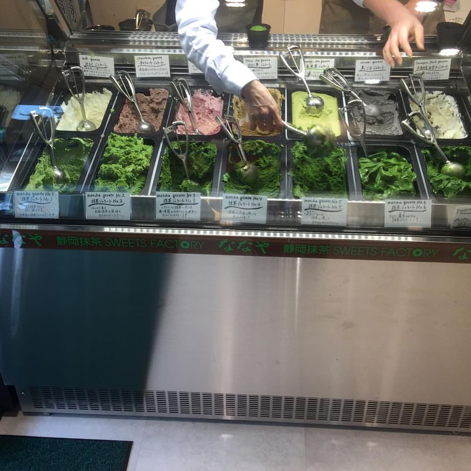 A chilled case of seven green matcha gelatos under hand-written flavour cards, a server's arm reaching in with a scoop