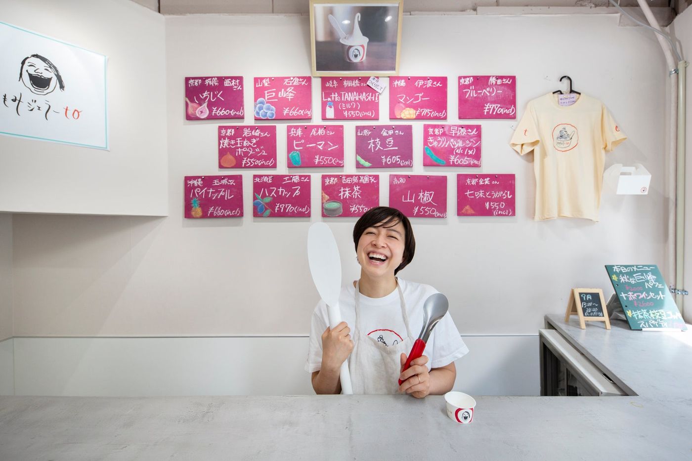 A woman in a white t-shirt and beige apron laughs behind a concrete counter holding a white spatula and a red-handled scoop, under sixteen handwritten magenta flavour cards and a sign with a laughing-face logo