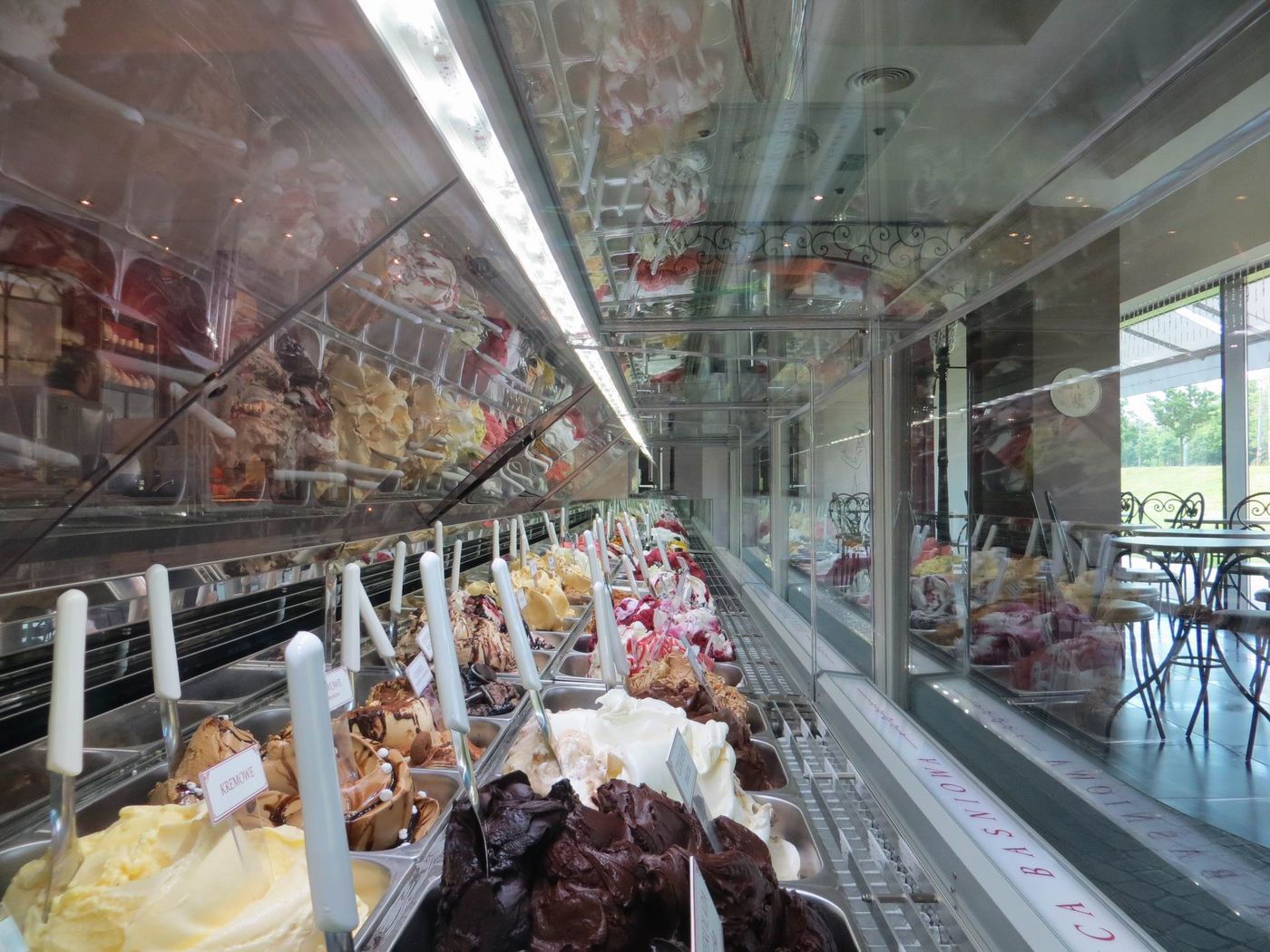 A gelato case runs into the distance packed with mounded tubs and long-handled spades, with BAŚNIOWA readable in reverse on the glass at right and metal café chairs and a window beyond