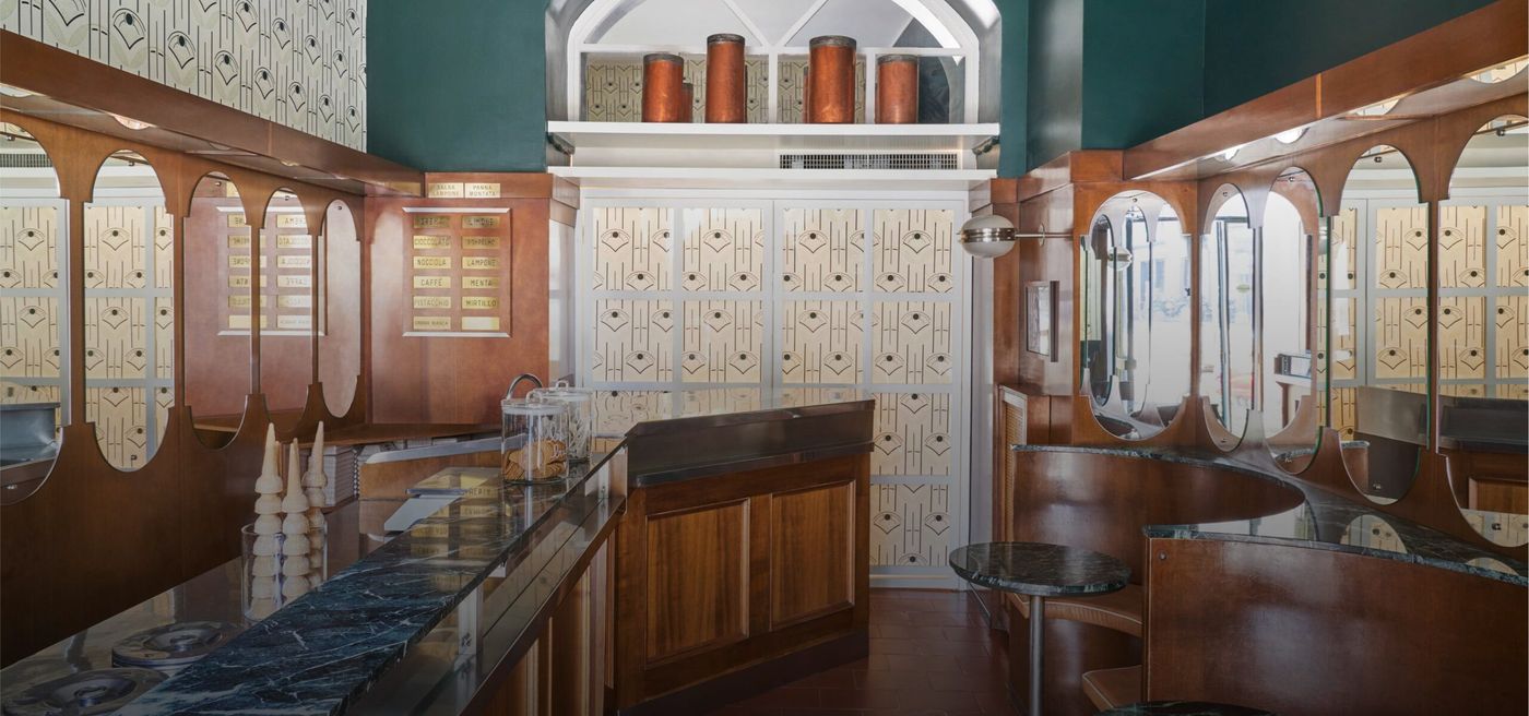 An Art Deco interior with pozzetti lids set into a green marble counter, brass-lettered flavour plaques reading CREMA, CIOCCOLATO, NOCCIOLA, CAFFÈ, PISTACCHIO, LIMONE, LAMPONE, MENTA and MIRTILLO, copper canisters on a high shelf and a glass of cones on the bar