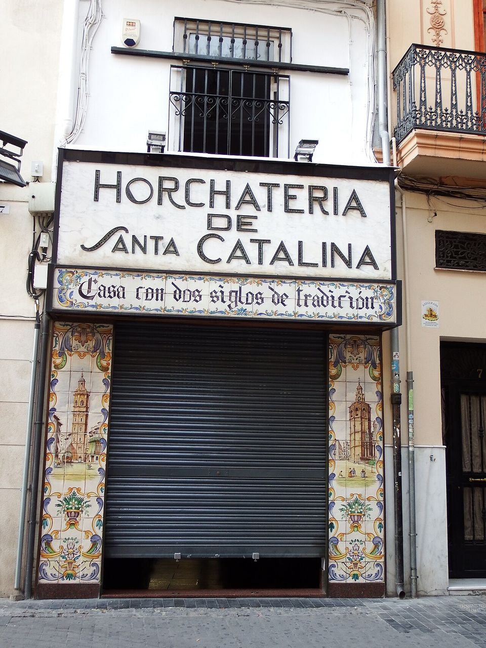 A marble fascia lettered HORCHATERIA DE SANTA CATALINA over a tiled panel reading Casa con dos siglos de tradición, painted bell towers flanking the shutter