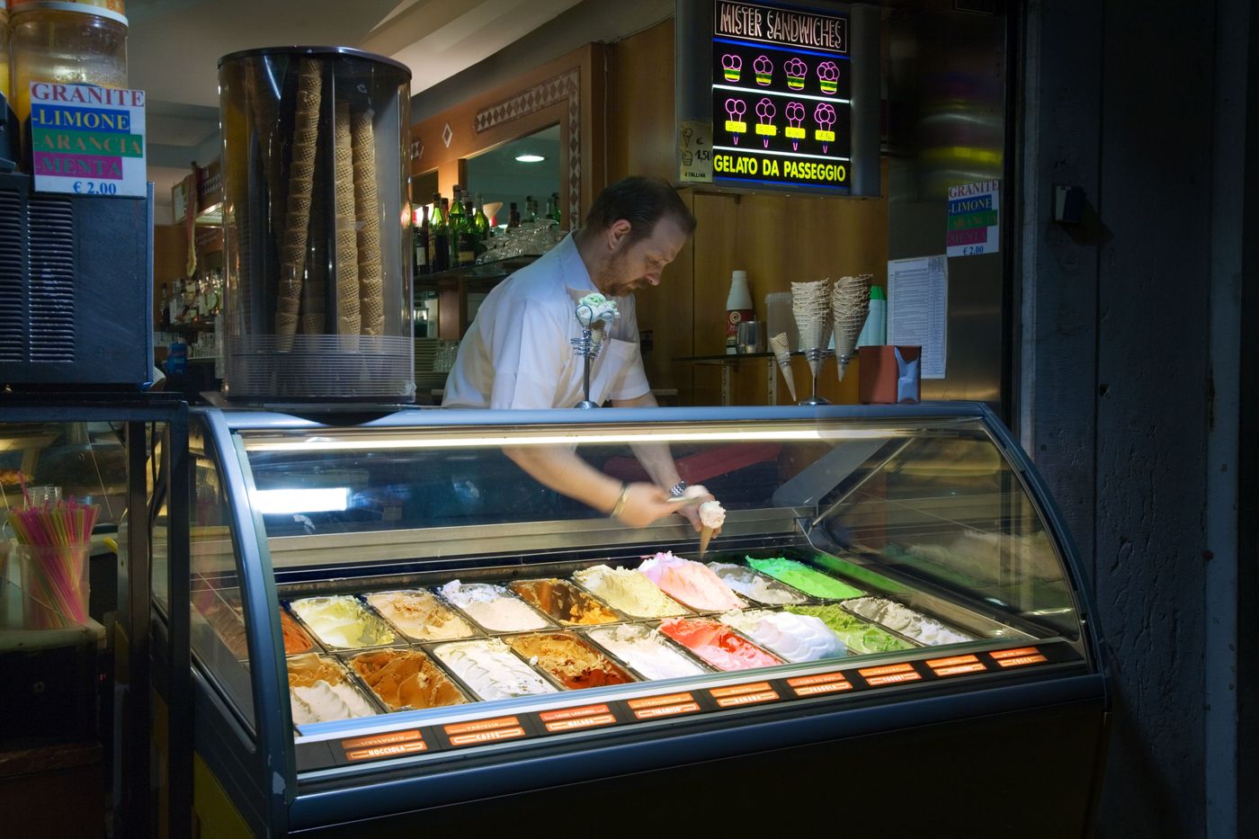 A man in a bow tie serving from a gelato case beneath a sign reading Gelato da Passeggio