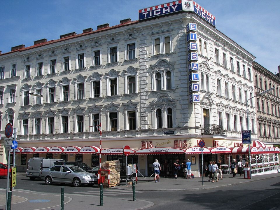 A corner Eissalon under red-and-white awnings lettered EIS, a TICHY sign on the roof and EISSALON running vertically down the façade, people at the door