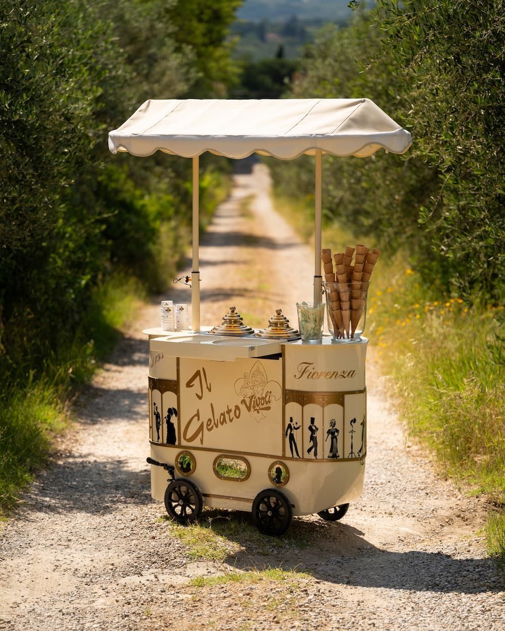 A cream-coloured gelato cart lettered Il Gelato Vivoli, standing alone on a gravel lane between hedges
