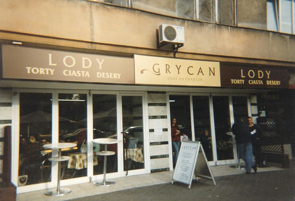 A shopfront signed GRYCAN between two boards reading LODY TORTY CIASTA DESERY, café tables visible through the glass