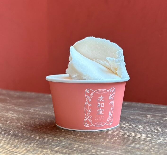 A single scoop of pale cream-coloured gelato in a pink cup branded Yuwado, Kyoto, against a dark red wall